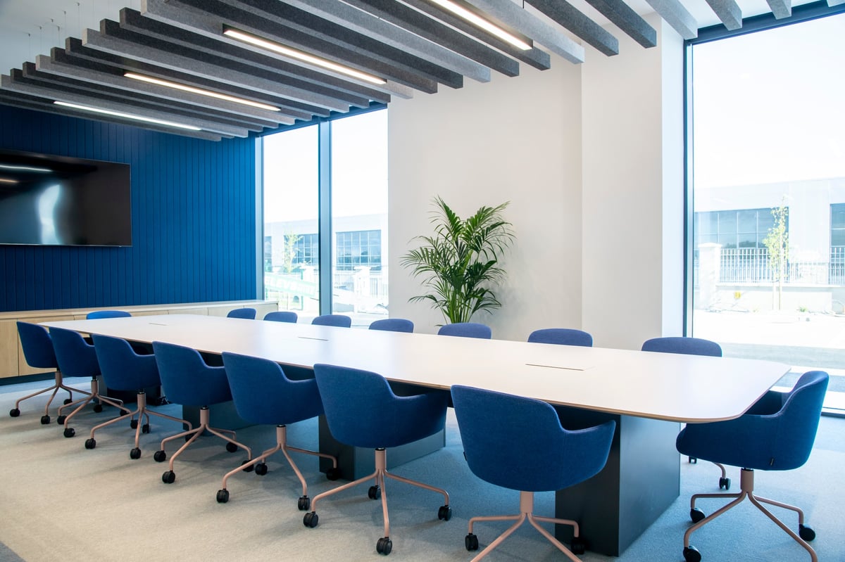 Modern conference room with long white table, blue chairs, geometric ceiling, large windows, and green plant
