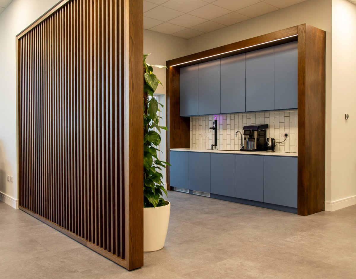 Modern office kitchenette with wooden slat divider wall, potted plant, and blue cabinetry behind glass doors