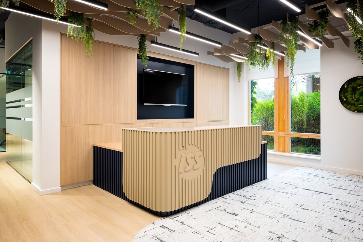 Modern office reception area with wooden and black corrugated desk, monitor display, hanging plants, and contemporary interior design