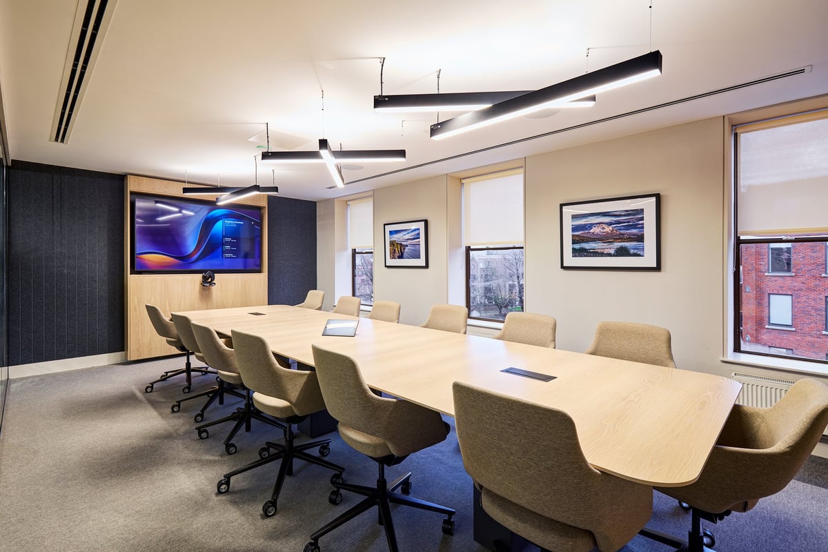 Modern corporate conference room with long white table, tan office chairs, wall-mounted TV, framed artwork, and contemporary pendant lighting