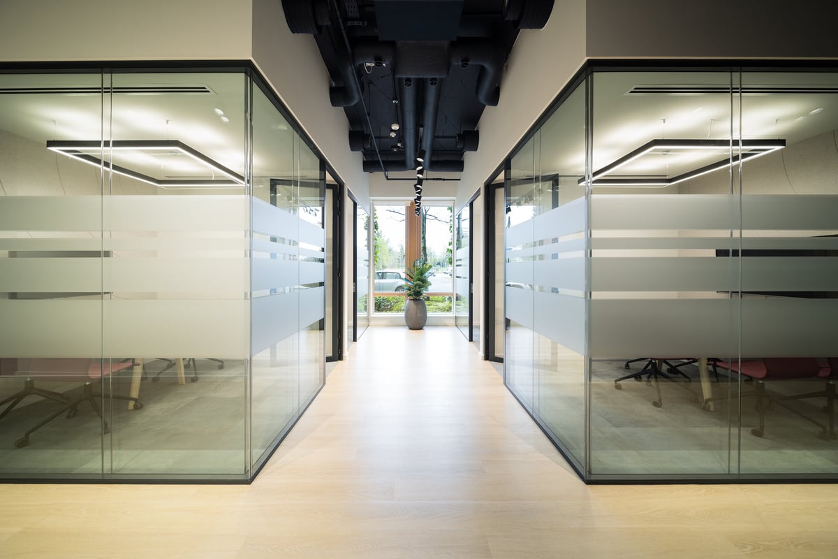 Modern office corridor with glass partition walls on both sides, frosted glass doors, and minimalist lighting fixtures on the ceiling
